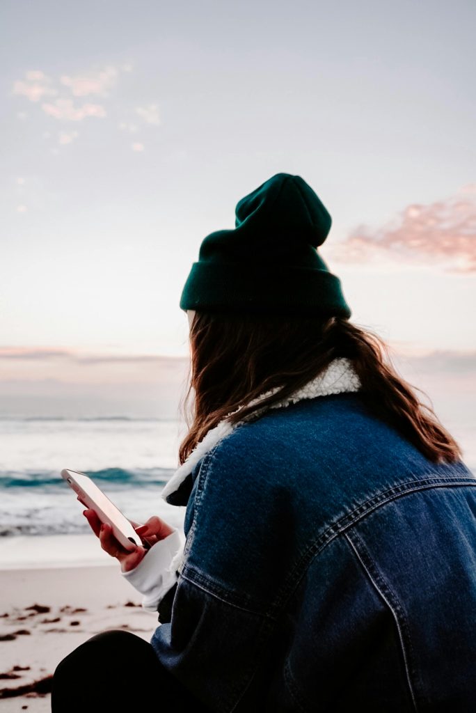 A woman in a beanie and denim jacket sits at a beach, holding a smartphone during a serene sunset.