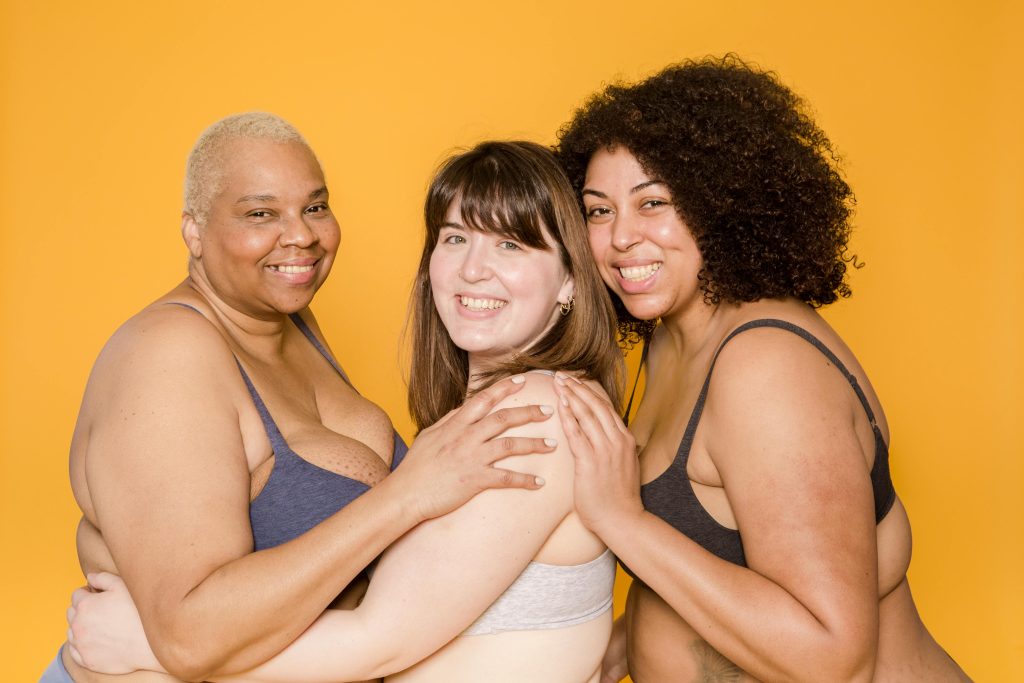Three diverse women embracing confidently, promoting body positivity against a yellow background.
