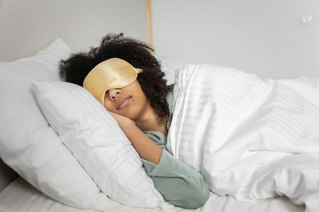 A woman with afro hair sleeps soundly in bed with a sleep mask, enjoying a cozy indoor atmosphere.