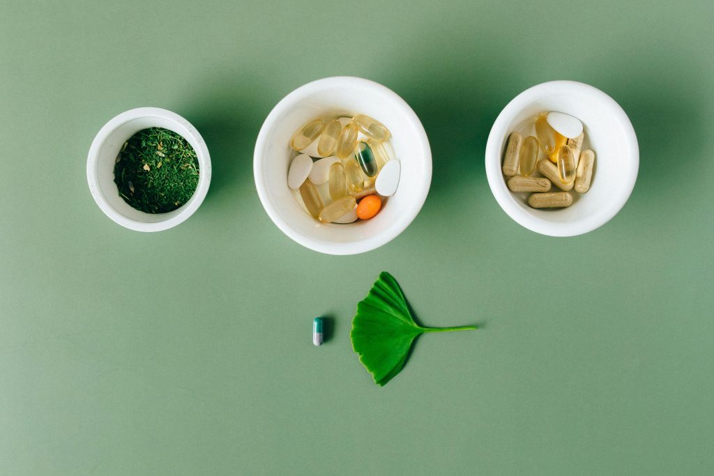 Flatlay of capsules and herbs in ceramic bowls on green surface.